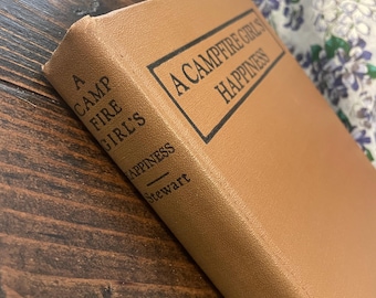 Op de afbeelding: Een boek met de titel "A Camp Fire Girls Happiness" met een bruine kaft. De titel staat in zwarte letters in een zwart kader. Het boek ligt op een donkerbruin houten oppervlak.