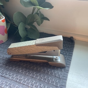 May include: A vintage stapler with a beige and gold finish rests on a gray textured surface. The stapler has a rectangular shape with a hinged top and a metal base. The text "LIFT COVER TO LOAD" is visible on the stapler.