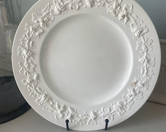May include: A round white ceramic plate featuring a raised relief border design of grapevines, leaves, and clusters of grapes. The center of the plate is smooth and plain. A small blue square icon with the white number 1 is visible.