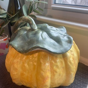 May include: A ceramic pumpkin-shaped container with a green leaf-shaped lid. The pumpkin is a warm yellow with orange accents, and the lid has a textured green finish. The container is likely used for storage or decoration.