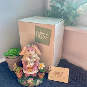 May include: A collectible figurine from the Cranberry Patch collection. The figurine features a pink bunny holding a smaller bunny, surrounded by potted flowers. The figurine is on a base with flowers and greenery.