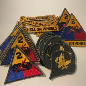 May include: A collection of military patches. Several triangular patches feature a yellow background with a black tank, the number "2", and the words "HELL ON WHEELS". Additional patches include a gold eagle design on a dark green background.