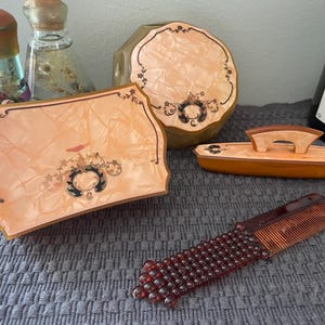 May include: A collection of vintage vanity items, including a rectangular tray, a round compact, and a small comb. The items are in shades of peach and brown, with decorative floral accents. A small wooden brush and two glass bottles are also present.