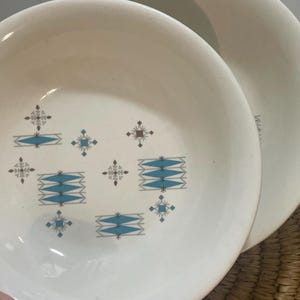 May include: Two off-white ceramic bowls featuring a repeating mid-century pattern of teal diamond shapes and brown starburst accents. The bowls have a smooth, rounded rim and a shallow basin, with the interior design centered on the base of each bowl.