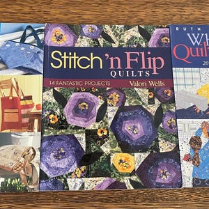 May include: Three books on quilting are displayed. The titles are "Patchwork with Pizzazz", "Stitch 'n Flip Quilts", and "When Quilters Gather". The covers feature images of quilts, bags, and people quilting. The books are on a wooden surface.