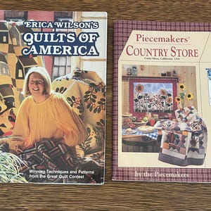 May include: Two books on a wooden surface. The book on the left is "Erica Wilson's Quilts of America" with a woman in a yellow shirt. The book on the right is "Piecemakers Country Store". Both books are about quilting.