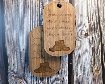 Könnte beinhalten: Zwei Holzhundemarken mit brauner Holzmaserung. Die obere Marke hat den Text "Some heroes wear capes. Mine wears combat boots." Die untere Marke hat den Text "Some wear capes. Mine wears combat boots." Beide Marken haben eine Silhouette eines Kampfstiefels.