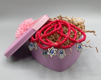 May include: A pink heart-shaped box filled with red beaded bracelets with silver hamsa hand charms and blue evil eye details. The box is filled with brown packing paper.