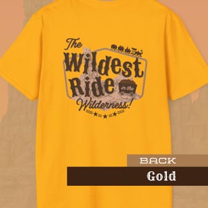 May include: Mustard-yellow t-shirt with the phrase "The Wildest Ride in the Wilderness!" in a vintage font. The design includes a mountain, a train, and a wagon. The back of the shirt is visible, with the word "Gold" at the bottom.