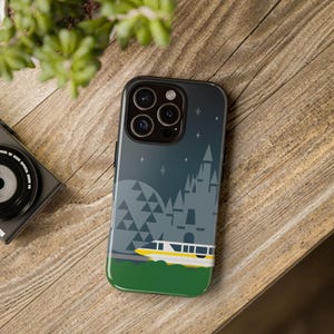 May include: A black phone case featuring a nighttime scene with a castle, geometric shapes, and a monorail. The case has a green base and a dark blue sky with stars. The phone case is on a wooden surface.
