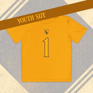 Pode incluir: Camiseta amarela mostarda para jovens com o número 1 em preto. Um pequeno brasão está acima do número. O texto "YOUTH SIZE" é impresso em uma faixa marrom na parte superior da camiseta.
