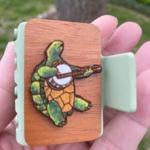May include: A wooden hair clip with a green and brown turtle playing a banjo. The clip is green and has a textured surface.