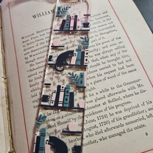 May include: A clear acrylic bookmark featuring a repeating design of bookshelves, cats, and teacups. The design includes teal, pink, and black elements. A pink ribbon is attached at the top. The bookmark is placed on an open book.