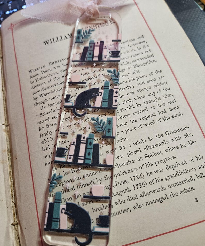 May include: A clear acrylic bookmark featuring a repeating design of bookshelves, cats, and teacups. The design includes teal, pink, and black elements. A pink ribbon is attached at the top. The bookmark is placed on an open book.