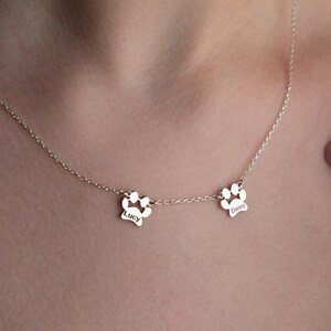 May include: A delicate silver necklace featuring two paw print pendants. Each pendant has a name engraved on it. The necklace is worn around the neck, showcasing the personalized design. The chain is thin and the pendants are small.