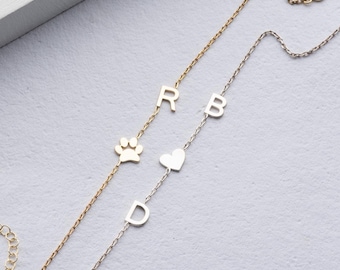 Puede incluir: Dos pulseras delicadas con dijes. Una pulsera es dorada con una huella de pata y la letra "R". La otra pulsera es plateada con un corazón y las letras "B" y "D".