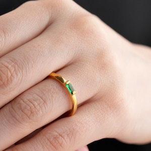 以下が含まれることがあります： 長方形のエメラルドグリーンの宝石が付いたゴールドリング。リングは指にあり、手は前景にあります。リングはシンプルでエレガントなデザインです。