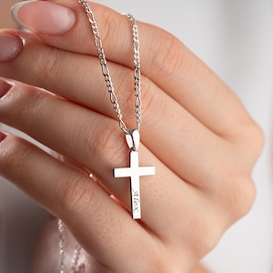Puede incluir: Un collar con colgante de cruz de plata con una cadena fina. La cruz tiene un diseño sencillo con el nombre "Alex" grabado. El collar se sostiene en una mano, mostrando los detalles de la joya.