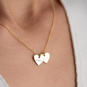 Op de afbeelding: Gouden ketting met twee hartvormige hangers. De linker hanger zegt "mama" en de rechter hanger zegt "forever".