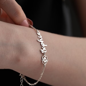 May include: A silver bracelet with the name "Sophia" in cursive script. The bracelet features a delicate flower charm and is being worn on a wrist. The background is dark, highlighting the jewelry.
