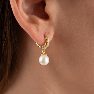 May include: Gold hoop earrings with a dangling pearl charm. The hoop earrings are adorned with small clear stones.