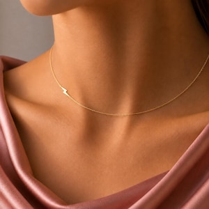 May include: A delicate gold chain necklace featuring a small, minimalist lightning bolt charm positioned off-centre. The thin, polished metal chain rests against the collarbone, showcasing a simple and refined design with a bright, reflective finish.