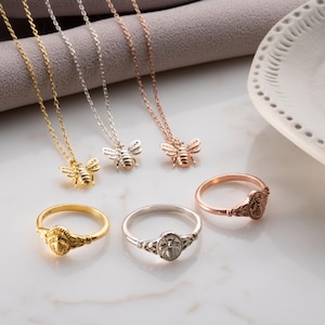 May include: Three necklaces with bee pendants and three matching rings in gold, silver, and rose gold tones. Each piece features a detailed bee motif. The necklaces have delicate chains, and the rings have a textured band design with an oval bee centerpiece.