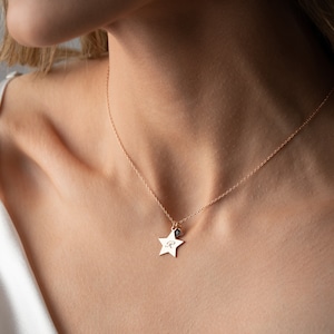 Puede incluir: Un delicado collar dorado con un colgante de estrella y un pequeño dije. El colgante de estrella tiene grabada la letra "R". El collar se lleva alrededor del cuello.