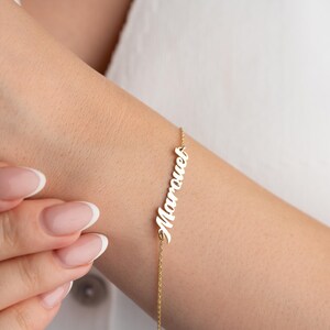 以下が含まれることがあります： 筆記体で「Marouel」という名前が入った繊細なゴールドブレスレット。ブレスレットは細いゴールドチェーンに付いています。ブレスレットは人の腕に着用されています。
