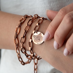 May include: A rose gold-toned multi-strand chain bracelet featuring a circular pendant engraved with a detailed cat portrait and the name REIS. The bracelet consists of alternating smooth oval links and twisted textured links in a metallic finish.