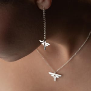 Puede incluir: Un conjunto de joyería en tono plateado que consta de un pendiente de hilo y un collar. Ambas piezas presentan un colgante simétrico y estilizado de polilla o luciérnaga con alas geométricas y un cuerpo central vertical, en cadenas finas.