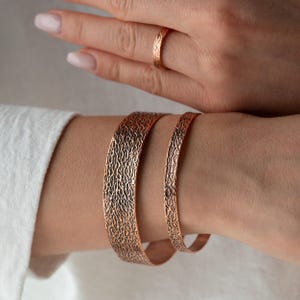 Op de afbeelding: Twee koperkleurige metalen armbanden met een donker, getextureerd, organisch patroon en een bijpassende dunne ring. De armbanden hebben een breed en een smal ontwerp, beide met een consistente, ruwe, geoxideerde afwerking op het oppervlak.