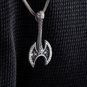 Könnte beinhalten: Eine silberne Axt-Anhänger-Kette mit filigranen Details. Der Axtkopf ist doppelseitig und hat ein keltisches Knotendesign.