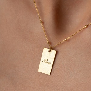 Könnte beinhalten: Eine goldene Kettenkette mit einem rechteckigen Anhänger in Form einer Plakette, auf der in kursiver Schrift "Rose" steht.