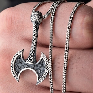 May include: A silver axe pendant with intricate details, hanging on a silver chain. The axe head has a curved blade and a detailed design on the surface. The chain is a box chain with a braided design.