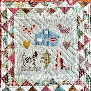 Op de afbeelding: Een patchworkdeken met een centraal paneel met een huis, een boom, een hond, een kat en twee vogels. De deken is gemaakt van verschillende stoffen in tinten roze, blauw, groen en bruin. De deken is omgeven door een rand van driehoeken in verschillende kleuren en patronen.