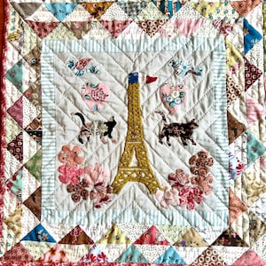Peut inclure: Une courtepointe blanche avec un motif de patchwork composé de tissus et de couleurs variés. Le centre de la courtepointe présente une tour Eiffel stylisée avec des chats et des cœurs. La courtepointe est entourée d'une bordure de petits carrés de patchwork.