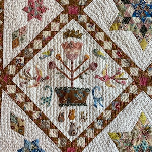 May include: Close-up of a handmade quilt with a repeating pattern. The quilt features a central floral design with birds and cats, surrounded by geometric shapes in various colors, including pink, blue, and brown. The background is white with a dotted texture.