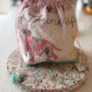 May include: A fabric drawstring pouch featuring a light pink background with a small floral pattern, a pink poodle graphic, and the embroidered word "Trinkets" in black thread. The bag includes two light pink drawstrings with small heart-shaped charms.