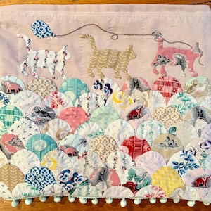 May include: A rectangular fabric wall hanging featuring a light pink background with three appliquéd cats connected by a stitched thread line. The lower section consists of overlapping scalloped fabric patches in various floral and geometric patterns.