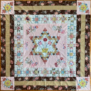 May include: A handmade quilt with a central star design featuring a floral arrangement in a yellow pot. The quilt has a pink centre, surrounded by brown floral and striped borders, and floral corner blocks. The quilt is a patchwork of various fabrics.