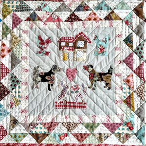 Puede incluir: Una tela acolchada blanca con un diseño de patchwork que presenta una casa, dos perros, dos pollos y un corazón. La tela está rodeada por un borde de triángulos en varios colores y patrones.