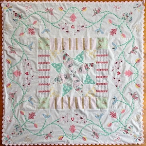 May include: A square quilt featuring a central star pattern made of floral and polka dot fabric patches, surrounded by a border of striped fabric. The outer white background displays green vines, pink animal figures, and a scalloped pink edge.