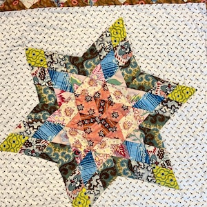 May include: A handmade quilt with a large star design in the center. The star is composed of various fabric scraps in shades of blue, pink, yellow, and brown. The background is white with a small, repeating pattern. The quilt's border features a geometric design.