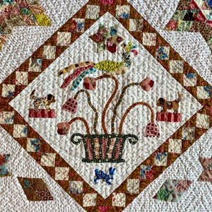 Könnte beinhalten: Nahaufnahme eines handgefertigten Quilts mit einem zentralen Rautenmuster. Der Quilt hat einen weißen Hintergrund mit einer floralen und Vogelapplikation. Das Design umfasst einen Vogel, Blumen und einen Korb, alles in verschiedenen Farben und Mustern. Der Quilt ist mit braunen Quadraten umrandet.