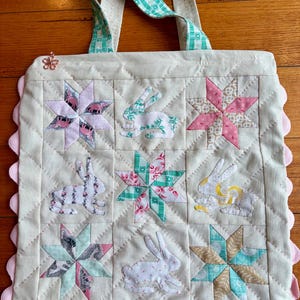 May include: A quilted tote bag featuring a grid of nine squares with alternating fabric appliques of rabbits and multi-coloured stars. The bag has two green and white checkered handles, a small flower-shaped zipper pull, and a scalloped pink trim.