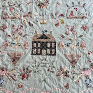 May include: A pale blue quilt with appliqued floral, butterfly, and house motifs. The central design features a house and the text "Hotel Bellineona 2035". Pink and white striped ribbon edges the quilt. The quilt is hand-stitched.
