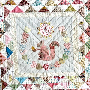 May include: A white quilt featuring a pink and brown squirrel design. The squirrel is encircled by a chain of hearts and a sun with a floral pattern. The quilt is crafted from patchwork fabric with a range of patterns and colours.