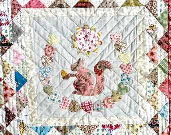 Könnte beinhalten: Eine weiße Steppdecke mit einem pinkfarbenen und braunen Eichhörnchen-Design. Das Eichhörnchen ist von einer Kette aus Herzen und einer Sonne mit einem Blumenmuster umgeben. Die Steppdecke besteht aus Patchwork-Stoff mit verschiedenen Mustern und Farben.