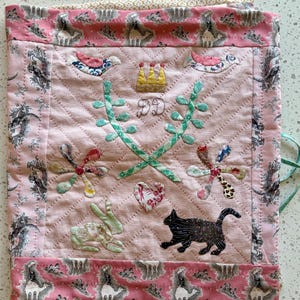 May include: A quilted fabric square featuring a light pink center panel with embroidered details including a yellow crown, the letters DD, two green leafy branches, a floral heart, a rabbit, and a black cat, framed by a pink border with camel motifs.
